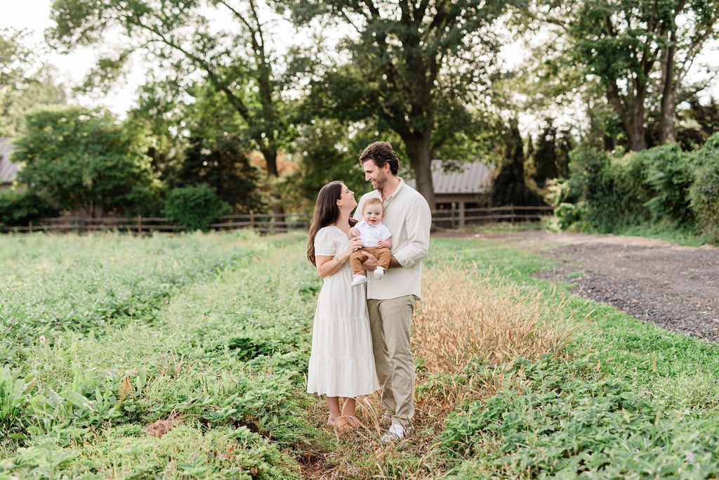 A mom, dad, and baby on a farm taking their photos together by a Philadelphia Family Photographer